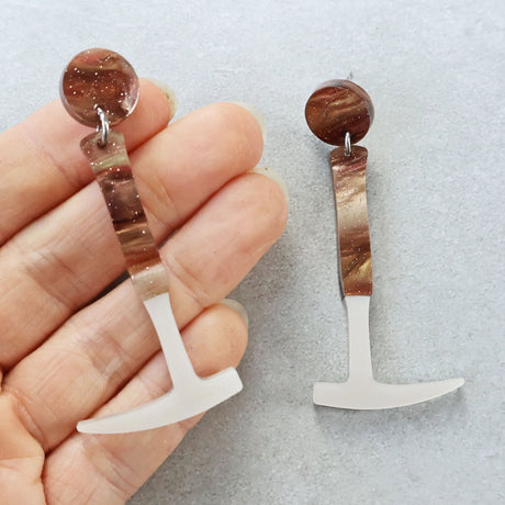 Laser cut acrylic fossil hammer shaped earrings being held up against a grey background. The dangly hammer handle and earring topper has brown swirls and glitter. 
