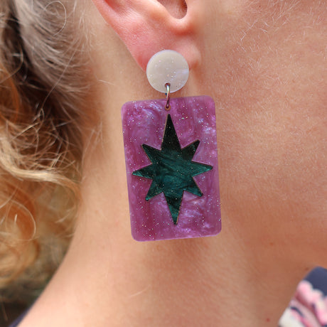 Earring with purple rectangular dangle and green star on a person's ear.