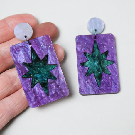 A pair of dangle earrings with a teal green star design set in a purple rectangle, and hanging from round earring toppers being held in a hand