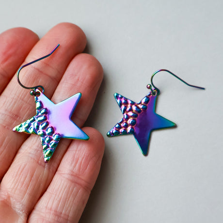 Star-shaped earrings with a metallic, iridescent finish held between fingers on a light background.