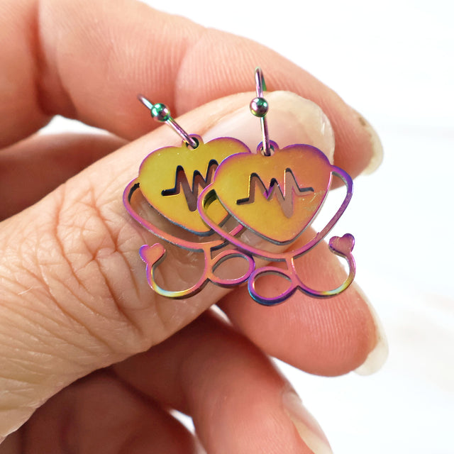 Stainless steel stethoscope earrings with french hooks, being held. Stainless steel is rainbow tinted. This particular pair has pink and yellow hues. 