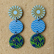 A pair of acrylic dangle earrings with circular designs in blue, green, and gold, showing sun, waves and seaweed graphics. 
