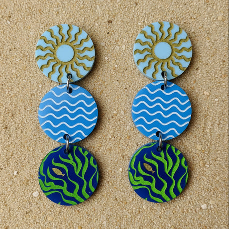 A pair of acrylic dangle earrings with circular designs in blue, green, and gold, showing sun, waves and seaweed graphics. 