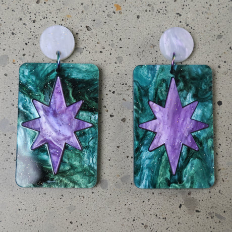 A pair of dangle earrings with a purple  star design set in a teal rectangle, and hanging from round earring toppers. 