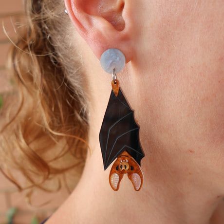Vampire bat-shaped acrylic earrings worn by a person. The bats hang upside down from a round earring topper. 