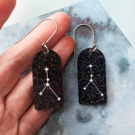 Black glittery arch shaped earrings with cancer star constellation design held by a hand on a textured surface.
