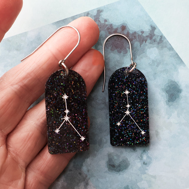 Black glittery arch shaped earrings with cancer star constellation design held by a hand on a textured surface.