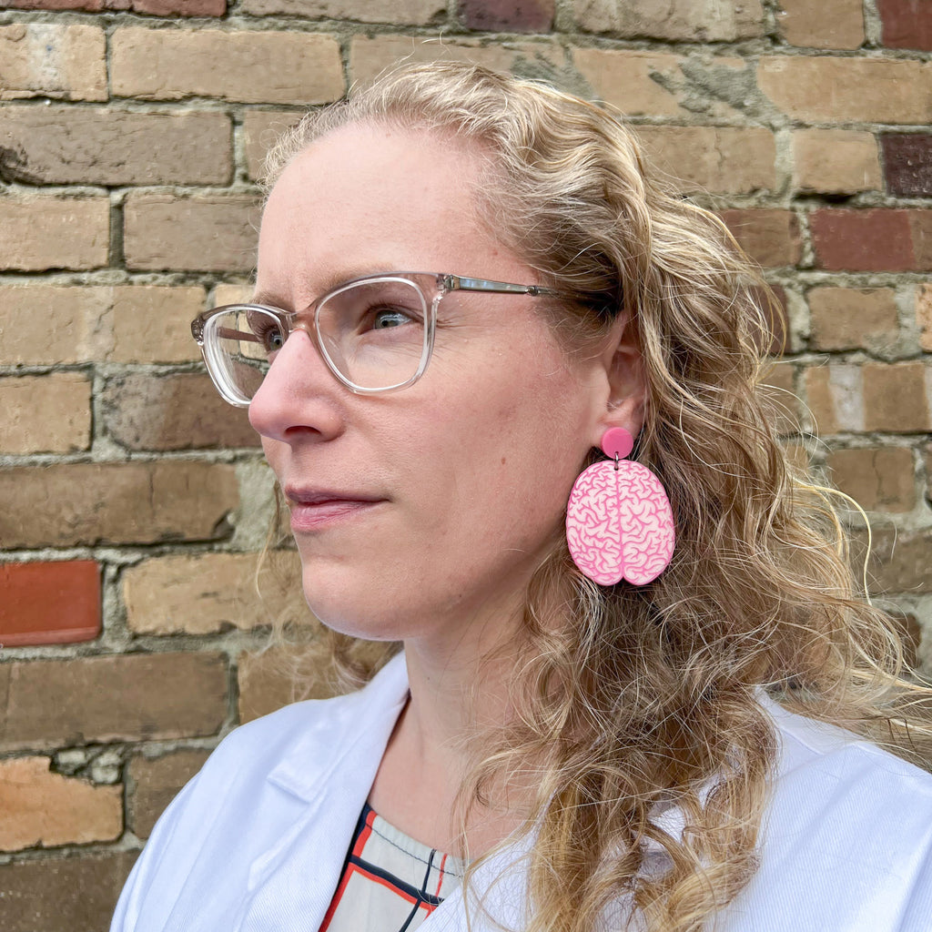 A pair of pink anatomical brain earrings being modelled by the maker, a blonde woman with glasses in a lab coat. Laser cut from acrylic.