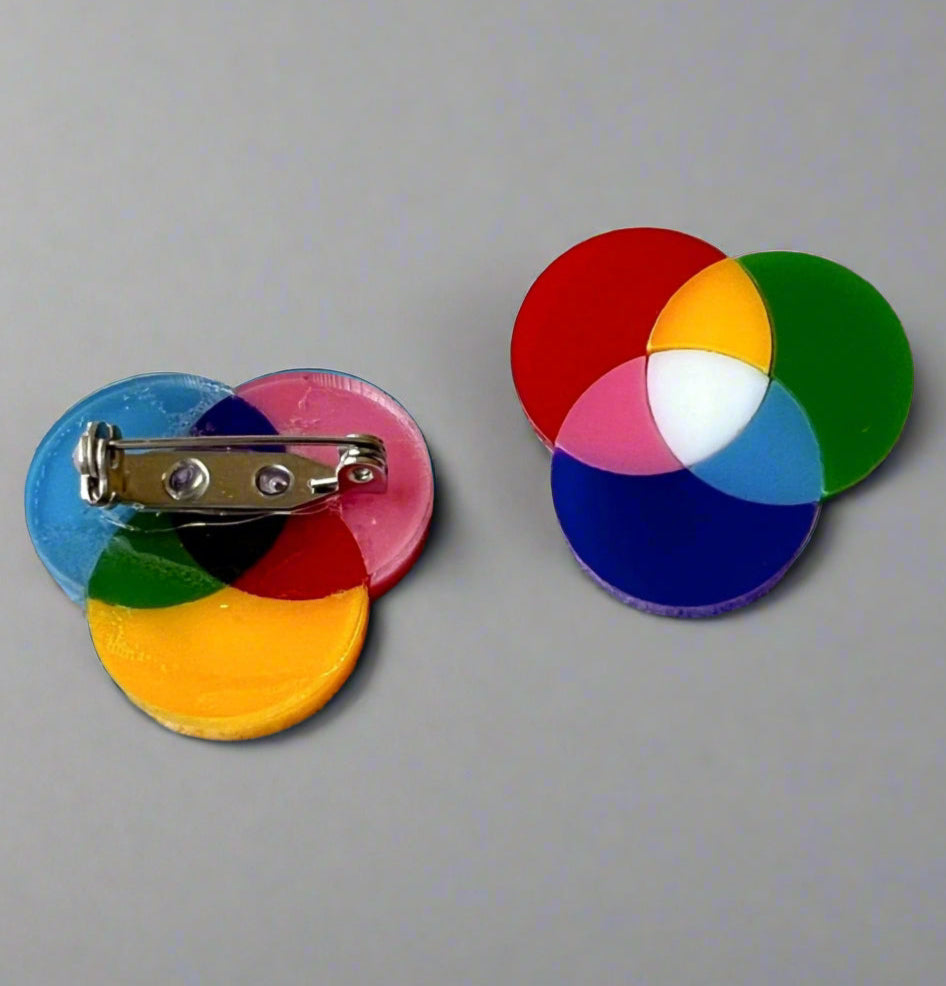 A pair of small subtractive and additive colour wheel brooches, laser cut in acrylic. The back of one brooch shows the stainless steel brooch pin with rolling clasp.