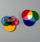 A pair of small subtractive and additive colour wheel brooches, laser cut in acrylic. The back of one brooch shows the stainless steel brooch pin with rolling clasp.