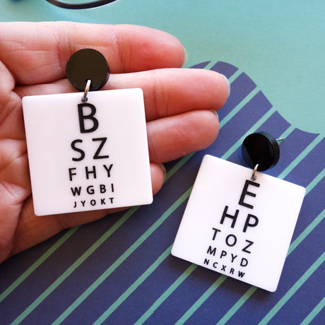 Laser cut acrylic eye chart earrings. With black text of a white background, and with black earring toppers. 
