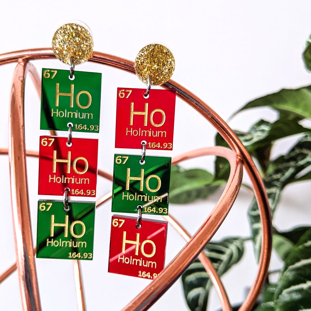 Ho Ho Ho drop earrings, featuring Holonium periodic table details and comprising of gold glitter acrylic, and transparent green and red acrylics, and gold painted lettering.