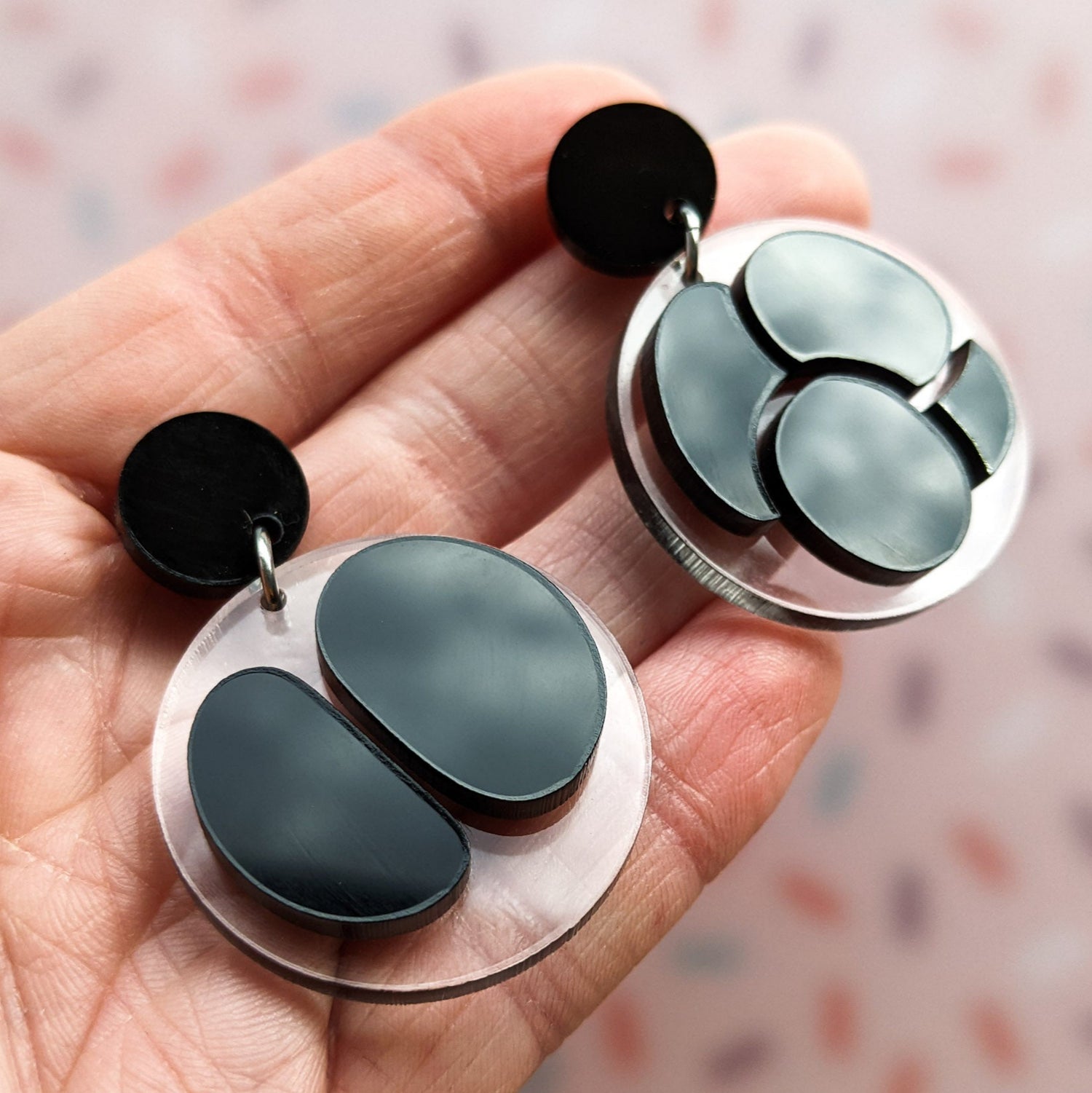 Asymmetrical meiosis earrings, with black shapes against clear circle backings, hanging from black earring toppers. Angled view in hands.