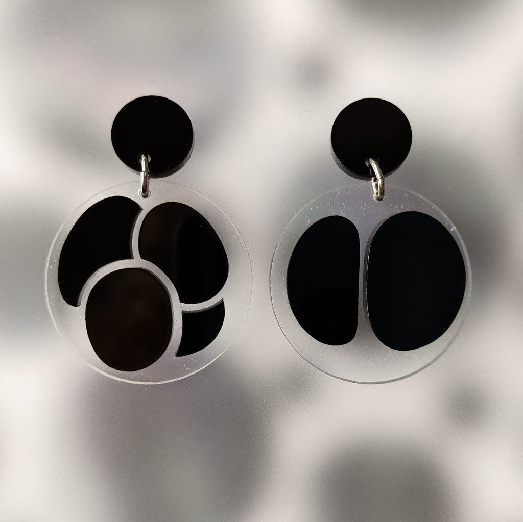 Asymmetrical meiosis earrings, with black shapes against clear circle backings, hanging from black earring toppers.