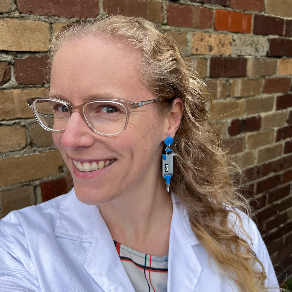 Grey and blue micropipette earrings laser cut and handpainted from acrylic. They hang from blue earring toppers. Earrings are being modelled by the maker, a blonde woman with glasses and a lab coat.