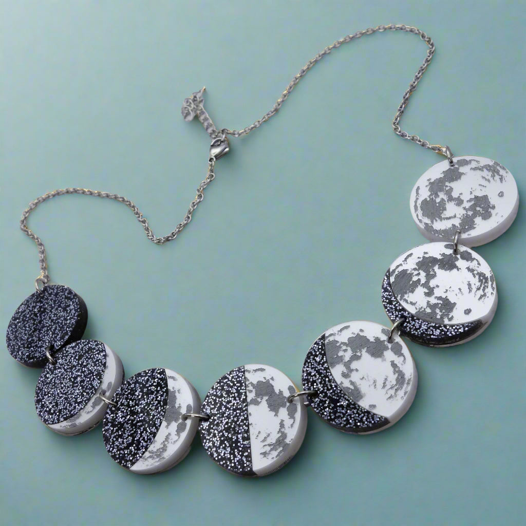 A laser cut acrylic moon phase necklace, showing the transition of the moon from new to full. Angled view.