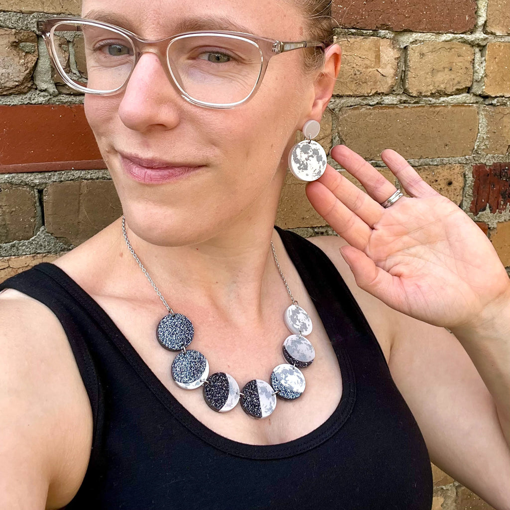 A woman modelling a pair of acrylic earrings and an acrylic moon phase necklace.