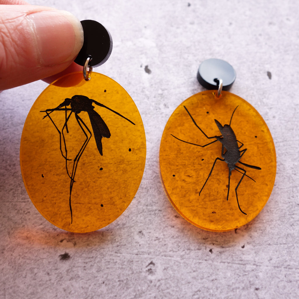 A pair of laser cut acrylic earrings, with black asymmetrical mosquito silhouettes engraved in transparent amber toned ovals.