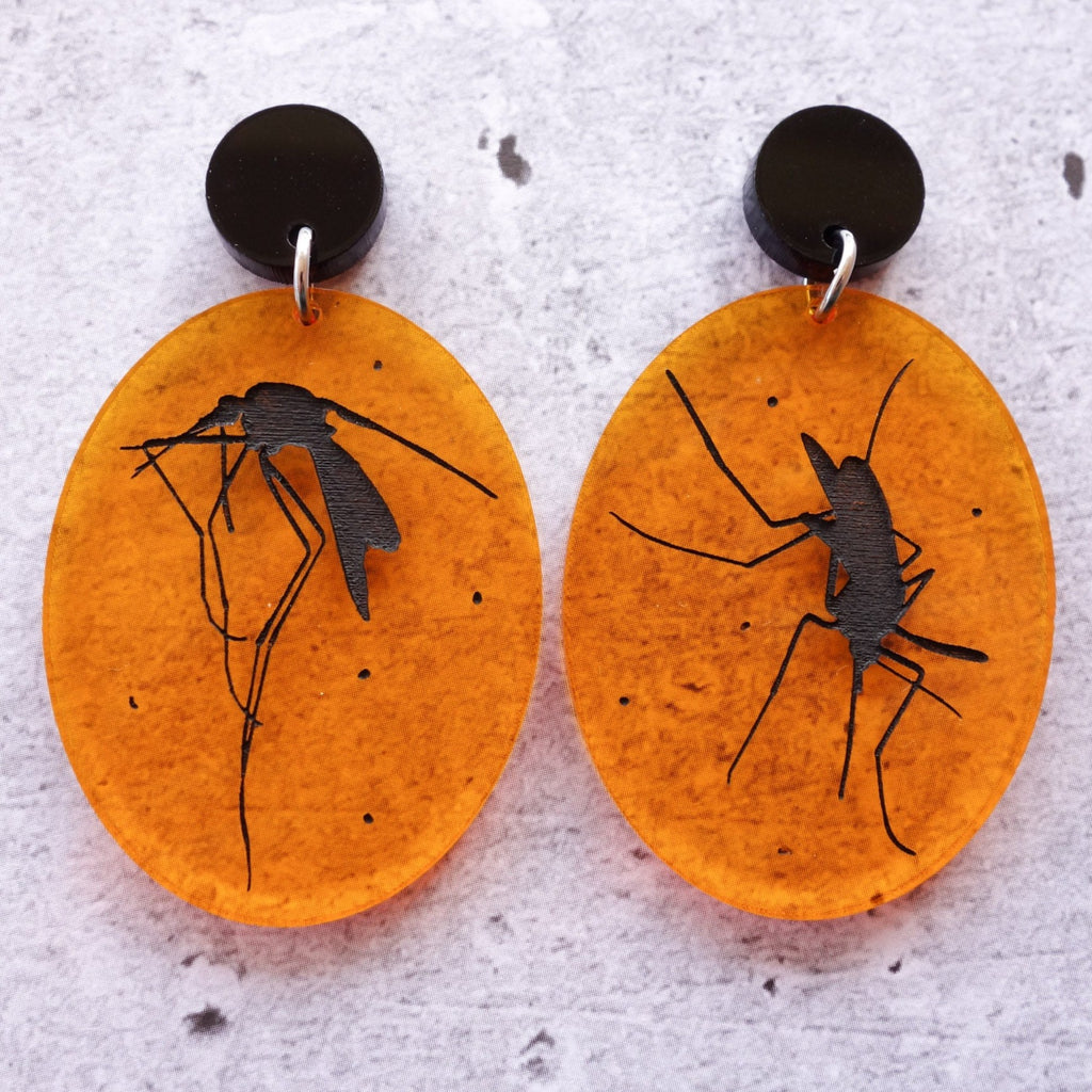 A closeup view of a pair of laser cut acrylic earrings, with black asymmetrical mosquito silhouettes engraved in transparent amber toned ovals. 