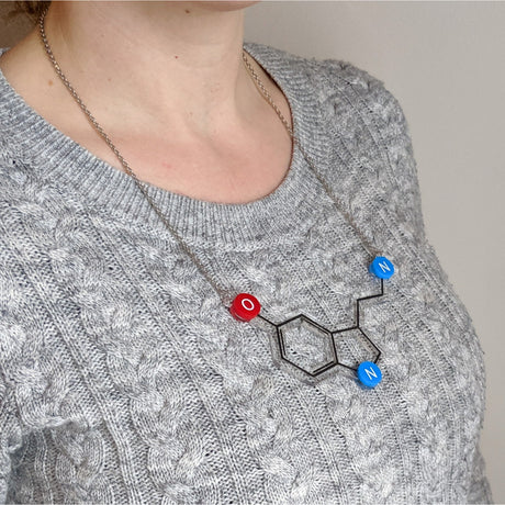 A person wearing a grey sweater is shown with a necklace featuring the serotonin molecule design. The necklace is composed of clear, red, and blue acrylic pieces representing the molecular structure, with black paint detailing the chemical bonds.