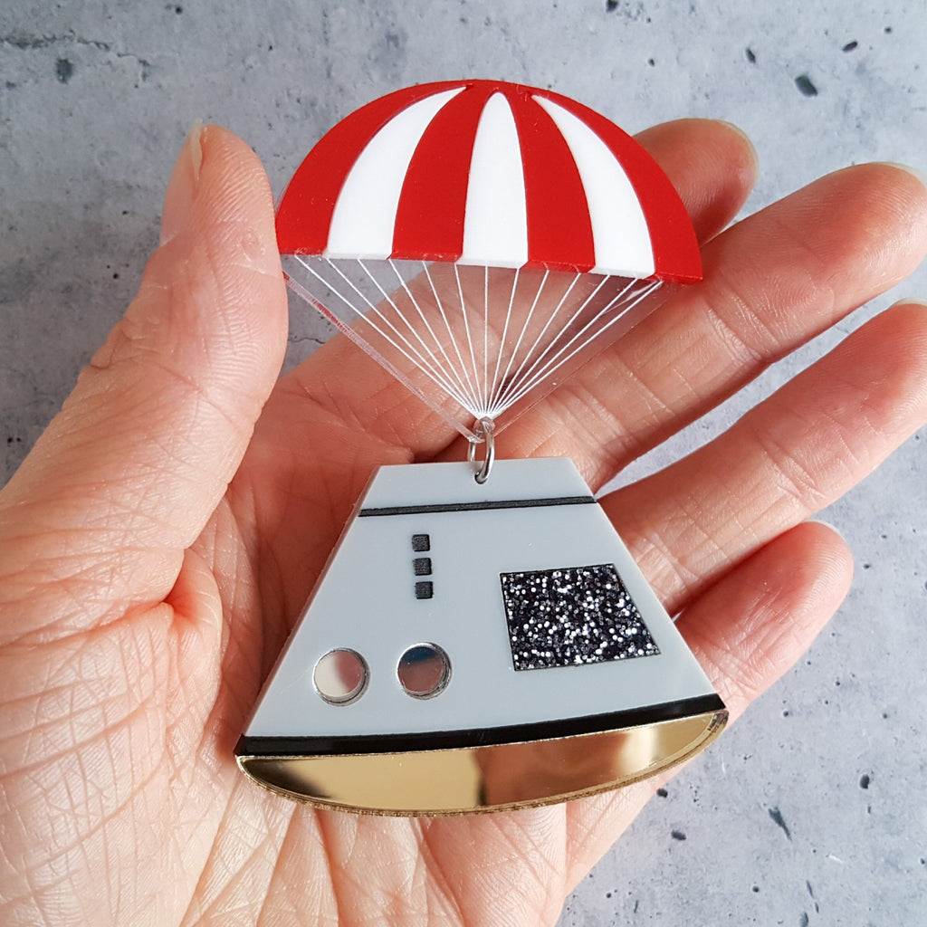 Space capsule brooch with red and white striped parachute, laser cut in acrylic. In hand for scale.