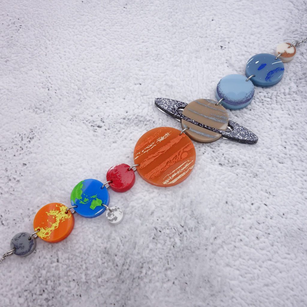 An angled view of a solar system necklace, showing Mercury, Venus, Earth, the moon, Mars, Jupiter, Saturn, Uranus, Neptune and Pluto in differently coloured acrylics with handpainted highlights.