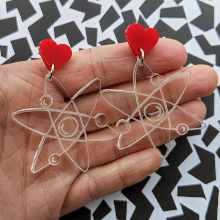 Atom earrings, laser engraved and cut from transparent acrylic, and hanging from red heart earring toppers.