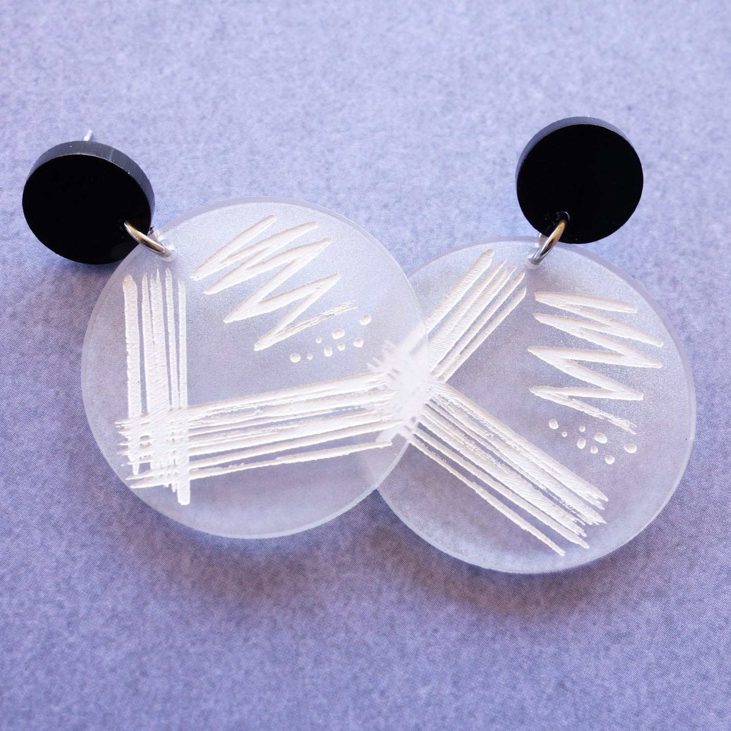 Streak plate earrings, with a streak plate pattern engraved and handpainted in white on a frosted clear acrylic disc. Earrings hang from black acrylic toppers. 