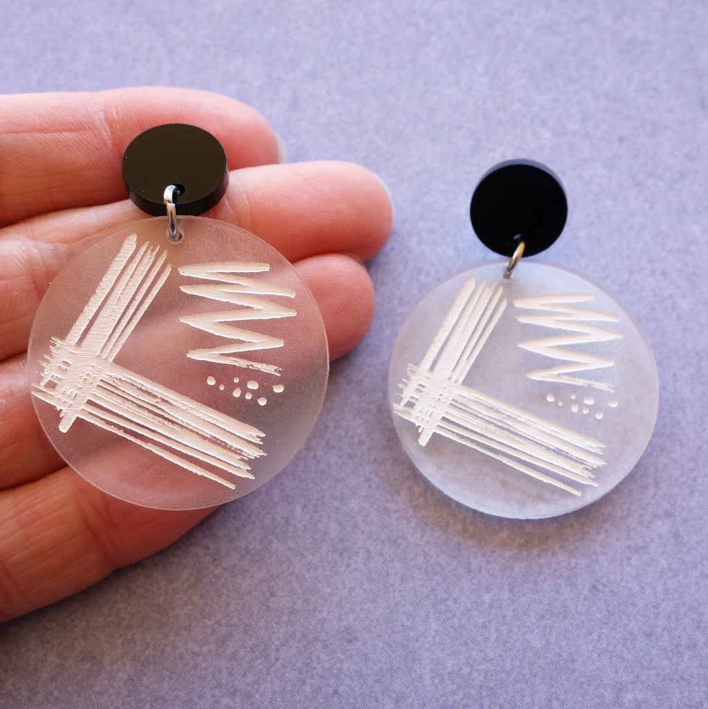 Streak plate earrings, with a streak plate pattern engraved and handpainted in white on a frosted clear acrylic disc. Earrings hang from black acrylic toppers. 