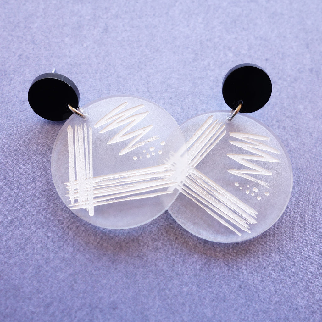 Streak plate earrings, with a streak plate pattern engraved and handpainted in white on a frosted clear acrylic disc. Earrings hang from black acrylic toppers. 