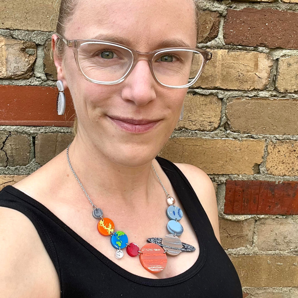 A solar system necklace, showing Mercury, Venus, Earth, the moon, Mars, Jupiter, Saturn, Uranus, Neptune and Pluto in differently coloured acrylics with handpainted highlights. Necklace is being modelled by the maker, a blonde woman with glasses.